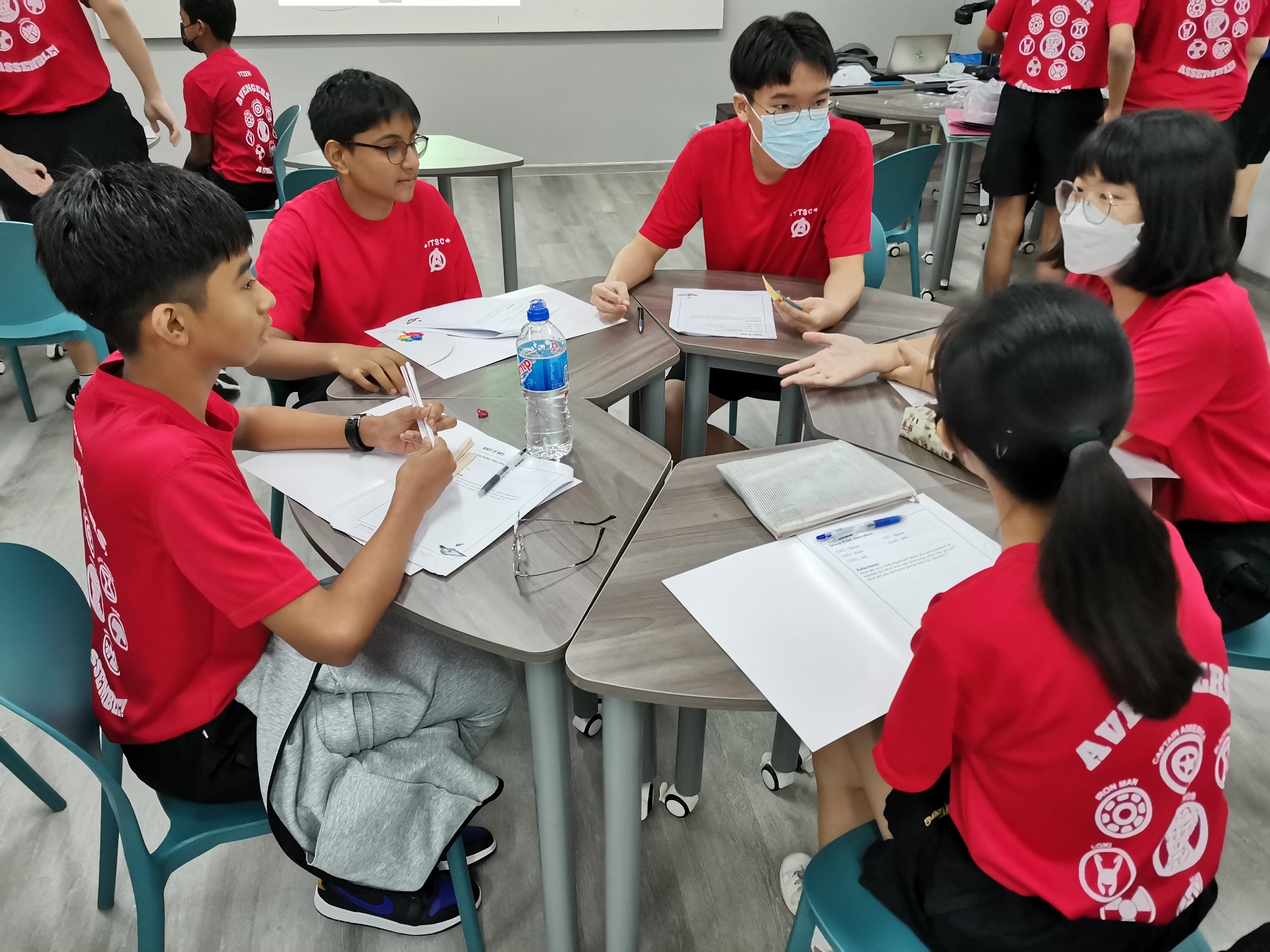 Students deep in discussion as part of the Aspiring Leaders programme.Five students in matching red shirts sit around joined desks with papers, discussing in a classroom setting.